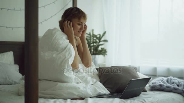Cheerful young woman using her laptop computer sitting in bed at home - Video