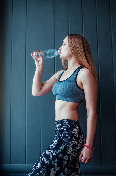 Shot of young woman drinking water at the gym. Muscular female having rest and drinks water in front of the window. - Photo, image