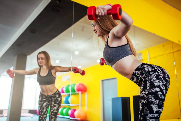 Снимок молодой женщины, тренирующейся с гантелями в спортзале. Мышечная женская тренировка перед зеркалом
. - Фото, изображение