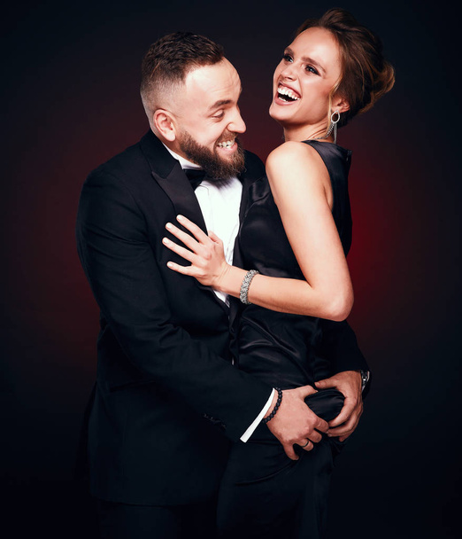 Luxurious, elegant and positive couple: handsome man with beard wearing tuxedo with amazing woman with blonde updo hair, wearing silky black dress and chic jewelry, posing in dark studio - Foto, Imagen