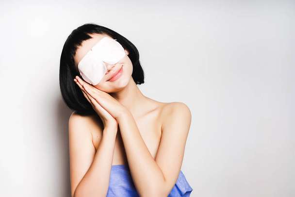 smiling young girl with black hair, in a blue towel, with a white mask before her eyes wants to sleep - Photo, Image