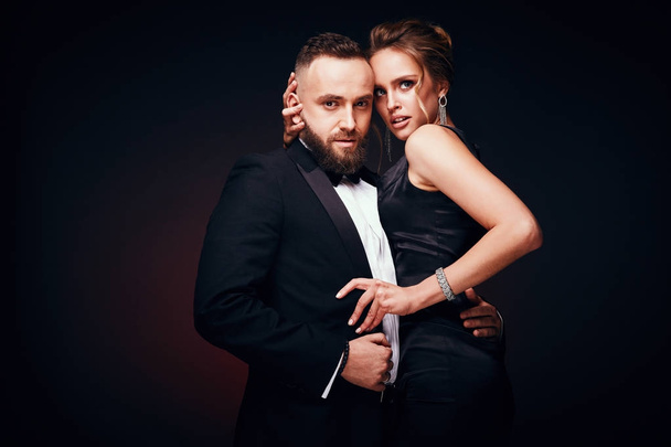 Passionate elegant couple: handsome bearded man in tuxedo with amazing woman with blonde updo hair, wearing silky black dress and chic jewelry, posing in dark studio - Photo, Image