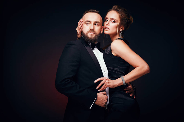 Passionate luxury couple: handsome bearded man in tuxedo with amazing woman with blonde updo hair, wearing silky black dress and chic jewelry, posing in dark studio - Foto, Bild