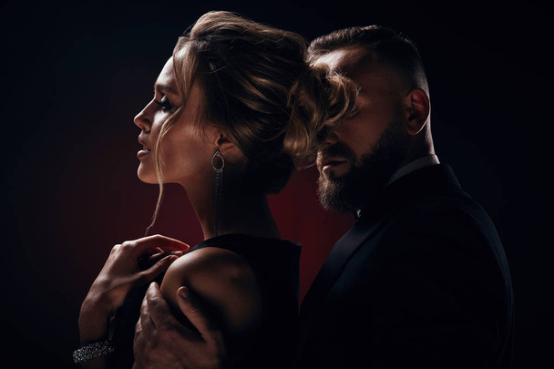 Luxurious woman with blonde updo hair, wearing silky black dress and chic jewelry and handsome bearded man in tuxedo with amazing, posing in dark studio - Foto, imagen