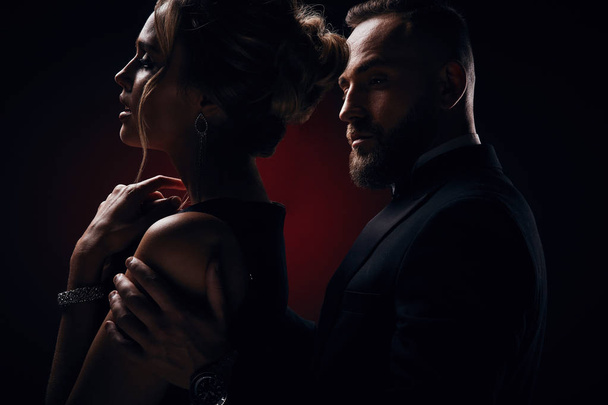 Passionate woman with blonde updo hair, wearing silky black dress and chic jewelry and handsome bearded man in tuxedo with amazing, posing in dark studio - Photo, Image