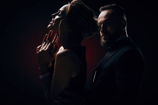 Sexy and luxury couple: woman with blonde updo hair, wearing silky black dress and chic jewelry and handsome bearded man in tuxedo with amazing, posing in dark studio - Zdjęcie, obraz