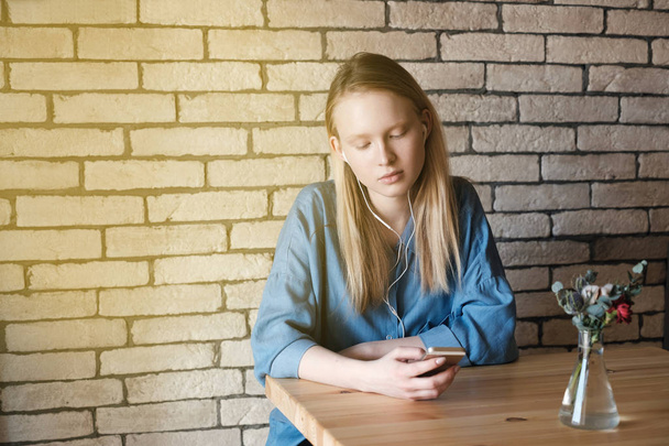 Sad blonde girl in blue blouse sitting at a table in a cafe, listening music with headphones and looking to the phone. - Photo, Image