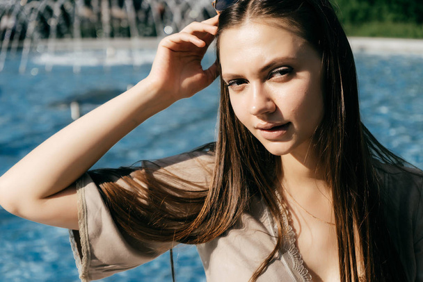 charming beautiful girl with long hair sitting by the pool with blue water, posing in the sun - Zdjęcie, obraz