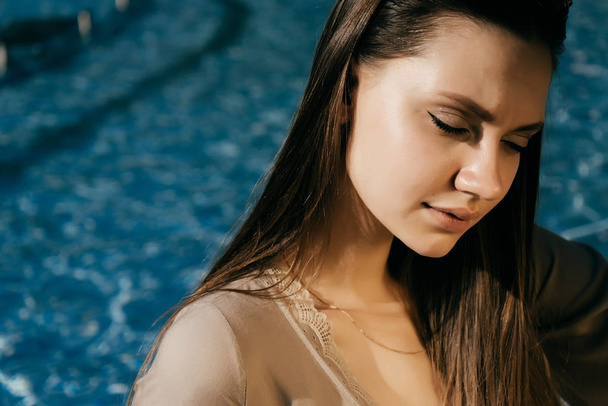 charming beautiful girl with long hair sitting by the pool with blue water, posing in the sun, her eyes closed - Fotoğraf, Görsel
