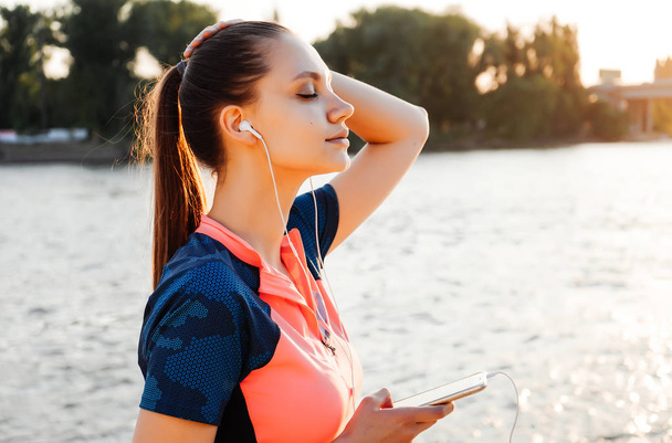 happy young fitness girl in sportswear listening to music on headphones, enjoying the sun by the river - Foto, imagen