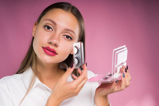 beautiful long-haired girl in a white shirt is holding a wrapped package with false eyelashes, on a pink background - 写真・画像