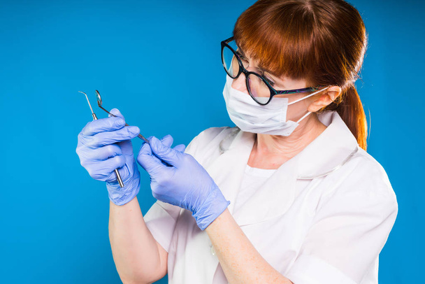 adult red-haired woman doctor in glasses with a protective mask and white coat holds in hands a medical dental instruments - Photo, image