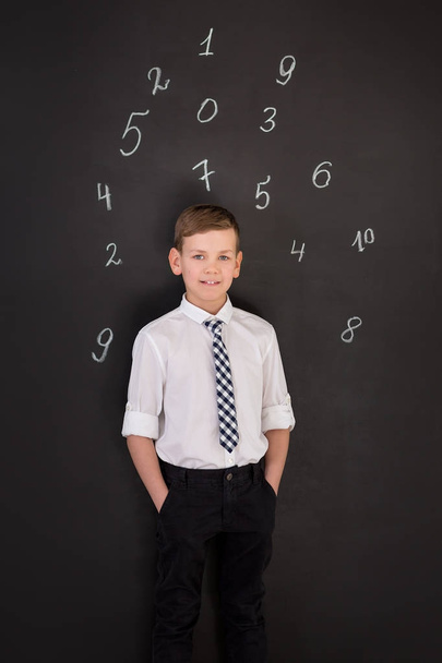 Симпатичный школьник в галстуке в белой рубашке стоит рядом со школьным столом с цифрами выше головы
. - Фото, изображение
