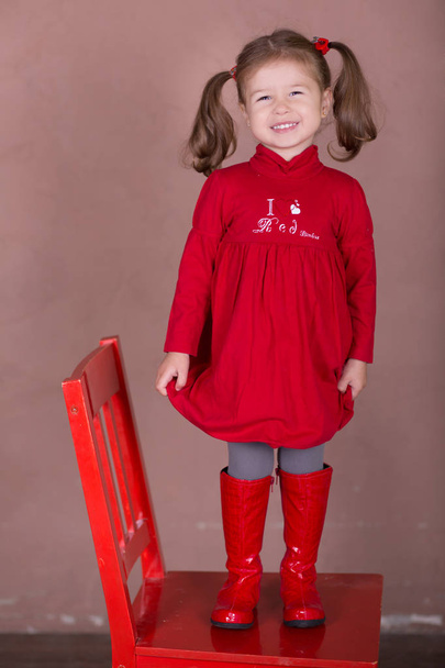 Mooi meisje met brunnete haar stijlvol gekleed rode lange shirt-jurk met cornrow happyly glimlachend poseren camera fotosessie in moderne showroom studio dragen baby laarzen vergadering staande op stoel. - Foto, afbeelding