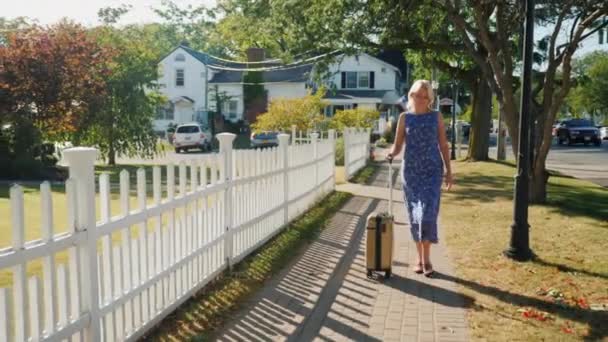 A woman with a travel bag on wheels is walking along the sidewalk. A typical American town. Steadicam shot - Záběry, video