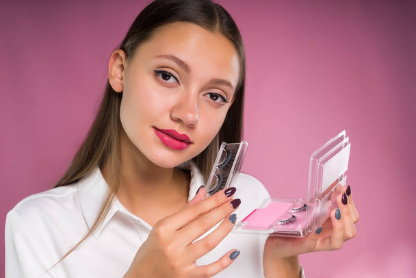 a beautiful girl with big lips takes out false eyelashes from the box and is going to stick them to herself and looks into the camera, isolated on a pink background - Фото, изображение