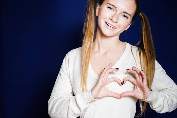 smiling girl in a white sweater with two ponytails shows her hands a heart and looks into the camera, isolated on a dark blue background - Valokuva, kuva