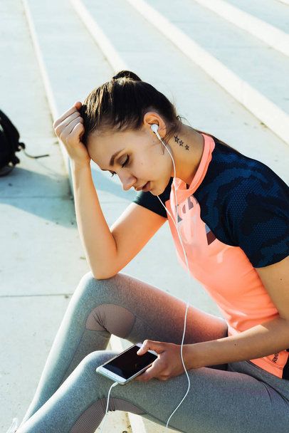 girl in a sport suit sits on the steps and listens to music on headphones - Φωτογραφία, εικόνα