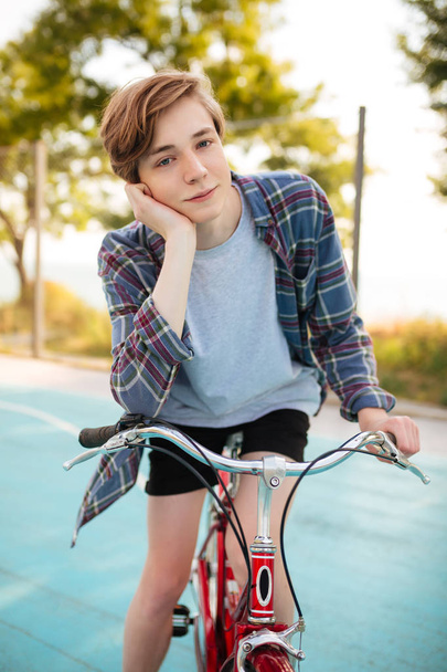ショート パンツでブロンドの髪を持つ少年、公園のバスケット ボール コートに赤い自転車でカジュアル シャツ立っての肖像画。若い男はうっとりと古典的な自転車で立ちながらカメラで探して - 写真・画像