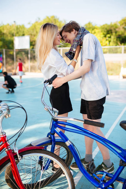 Miły chłopak i dziewczyna z blond włosami, spędzać czas razem i zabawy na boisko do koszykówki. Portret Młoda para stałego i szczęśliwie patrząc na siebie z niebieski i czerwony rower obok - Zdjęcie, obraz