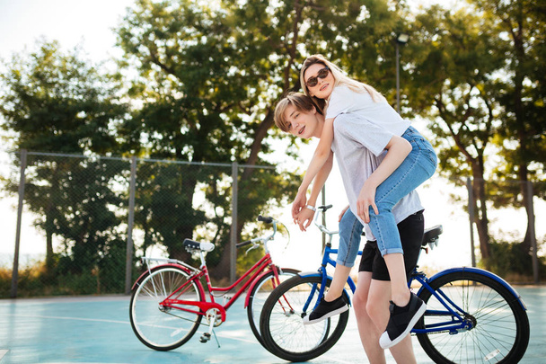 Portrait of young couple having fun in park with red and blue bicycle on background. Smiling boy looking in camera while playing with beautiful girl in sunglasses and holding her on his back  - Photo, Image