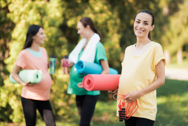 Three pregnant girls came to the park on fitness. A girl in a yellow T-shirt posing with a rug for yoga and a rope - Фото, изображение