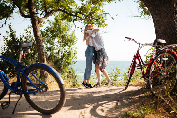 Портрет красивої пари, що обіймає один одного, стоячи в парку з дивовижним видом на море на фоні. Молода мила пара проводить час разом з червоними і синіми класичними велосипедами поруч
 - Фото, зображення