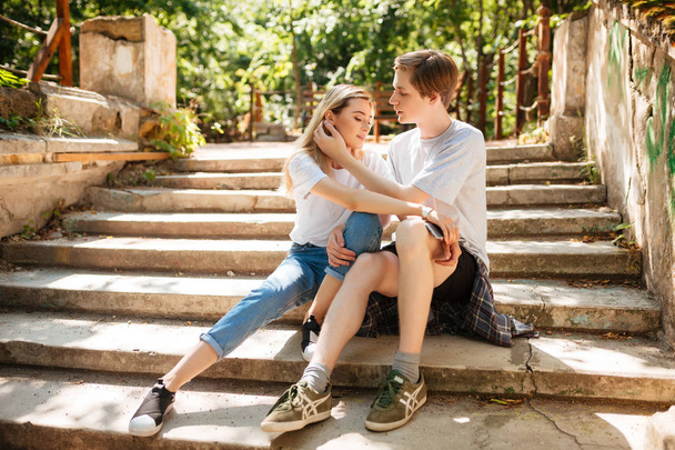 Young beautiful couple sitting on stairs in park and embracing one another while spending time together. Portrait of cool boy and girl with blond hair  sitting and thoughtfully looking aside in park - Photo, Image