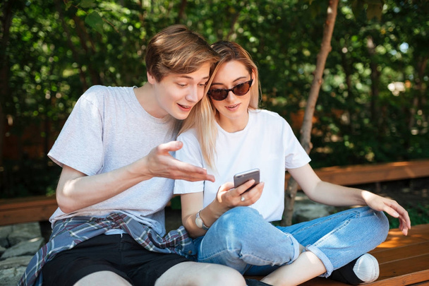 Portrait of joyful couple sitting on bench in park and emotionally discussion something while using cellphone. Close up photo of young smiling man and nice lady with blond hair spending time together  - Photo, Image