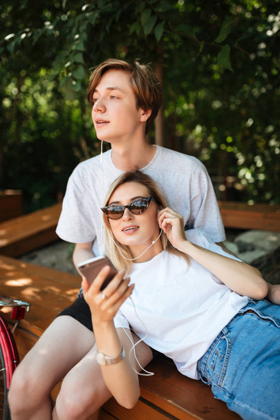 Portrait of young couple in earphones listening music while spending time in park. Thoughtful boy sitting on bench and dreamily looking eyes while beautiful girl leaning on him with cellphone in hand - Fotó, kép