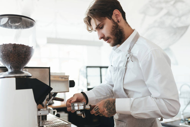 Portrait of young man working with coffee machine at the bar counter in coffee shop. Barista in apron and white shirt making coffee by coffee machine in restaurant. Cool boy working as barista at cafe - Foto, Imagen