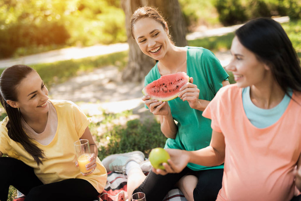 Tre donne incinte sedersi nel parco su un tappeto per pic-nic e mangiare. Stanno tutti sorridendo.
 - Foto, immagini