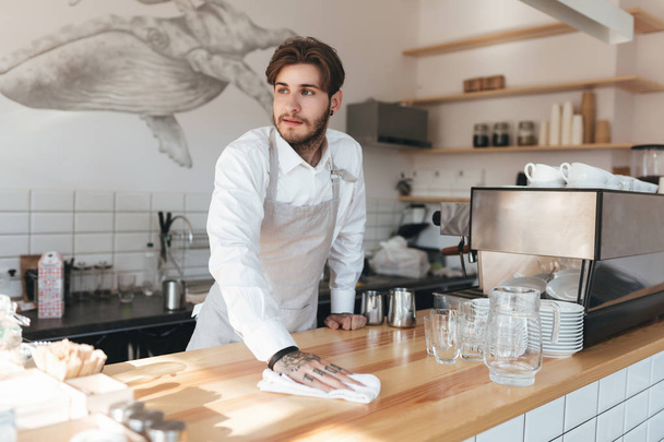 Young man thoughtfully looking aside and wiping counter at his workplace in restaurant. Portrait of barista in apron and white shirt standing at the counter in coffee shop - Foto, Bild
