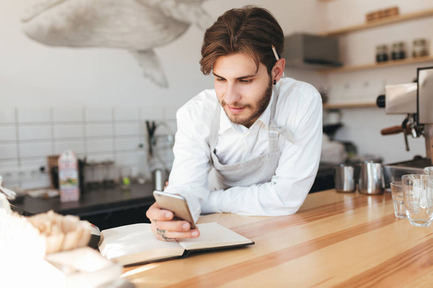 Portrait of young barista leaning his hands on counter happily looking in his mobile phone in hand at cafe. Boy in apron and white shirt with pencil behind ear standing at the counter in coffee shop - Foto, Bild