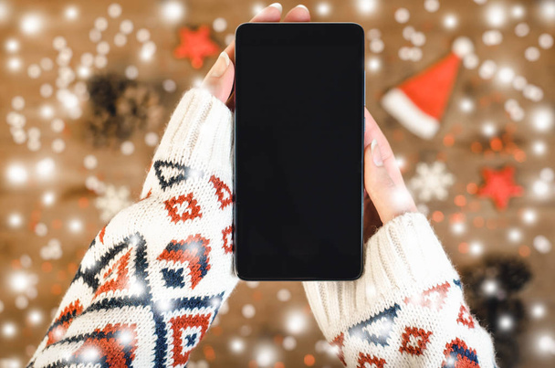 Manos femeninas en un suéter de punto cálido que sostiene el teléfono móvil inteligente con pantalla oled sobre fondo de madera con copos de nieve de Navidad y nieve. Feliz Año Nuevo y Navidad plano laico composición vista superior
. - Foto, Imagen