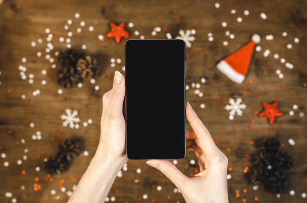 Manos femeninas en un suéter de punto cálido que sostiene el teléfono móvil inteligente con pantalla oled sobre fondo de madera con regalos de Navidad copos de nieve y nieve. Feliz Año Nuevo y Navidad plano laico composición vista superior
. - Foto, imagen