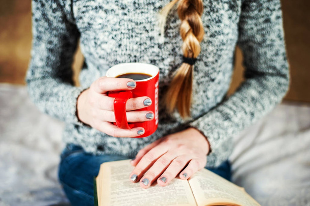 Молодая девушка в джинсах и свитере читает книгу с большой чашкой кофе или чая
. - Фото, изображение