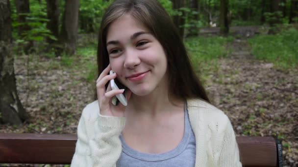 Young attractive girl speak to phone on bench. Summer park. Smile. HD video footage shooting static camera. - Felvétel, videó