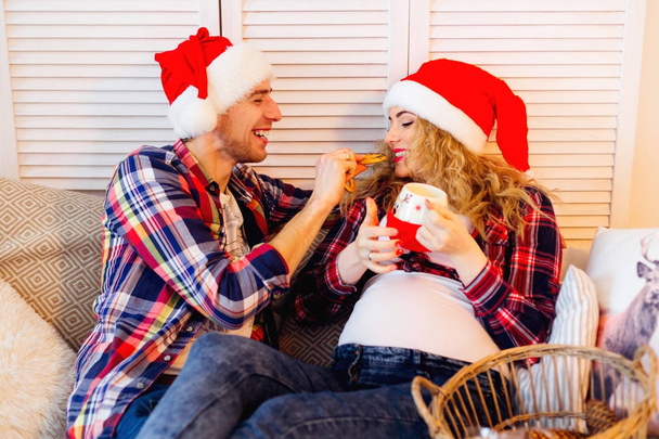 Noel Baba şapkalı bir adam onun hamile eşi kanepede beslenir. - Fotoğraf, Görsel