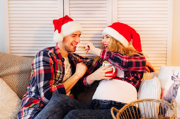 pregnant woman in Santa Clause hat feeds her husband - Photo, Image