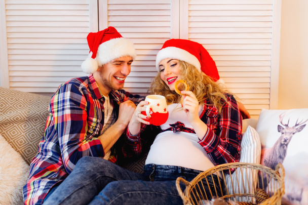 zukünftige Eltern im Hut des Weihnachtsmannes auf dem Sofa beim Plätzchenessen - Foto, Bild