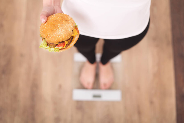 Ruokavalio ja pikaruoka käsite. Ylipainoinen nainen seisoo Punnitus Scale Holding Burger (Hamburger). Epäterveellistä roskaruokaa. Laihduttaminen, elämäntapa. Painonpudotus. Liikalihavuus. Ylhäältä
 - Valokuva, kuva