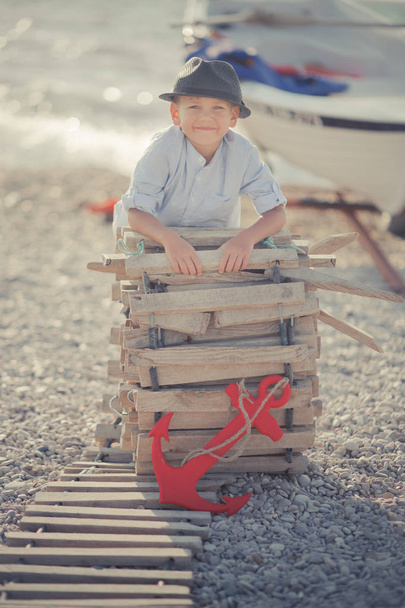 Handsome Nice looking boy on beach walking posing on wooden way wearing fancy stylish blue shirt and shorts with gallows and gentleman hat enjoy summer time alone on awesome ocean with anchor - Fotó, kép