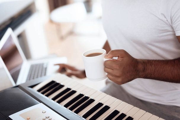 A black man sits in the living room of his apartment and plays a synthesizer. He composes music and drinks coffee. - Foto, imagen