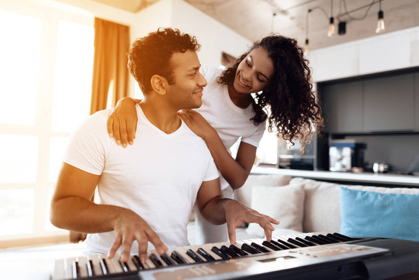 A black man sits in the living room of his apartment and plays a synthesizer. Nearby is his girlfriend and hugs him. - Photo, Image