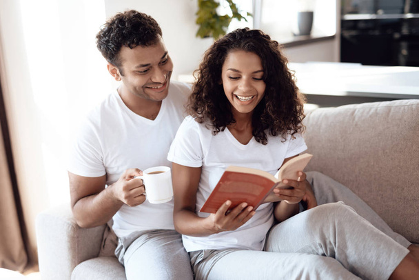 Un homme et une femme noirs sont assis sur le canapé. Une femme lit un livre, un homme est assis et la serre dans ses bras
. - Photo, image