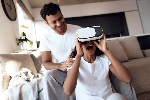 Black man and woman are sitting on the couch. The woman put on her helmet a virtual reality. - Foto, Imagen