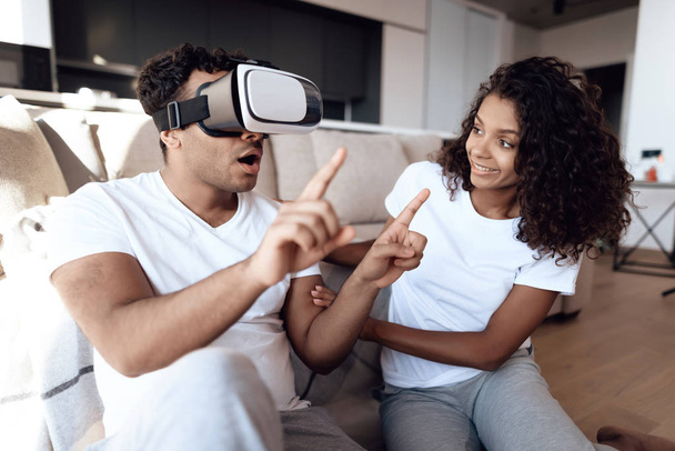 Black man and woman are sitting on the couch. The man put a vr helmet over his head and watches a thrilling film. - Zdjęcie, obraz