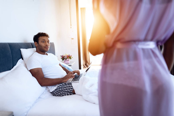 Black man and woman in the bedroom. A man is lying on the bed with laptop. Before him is his girl in sexy lingerie. - 写真・画像