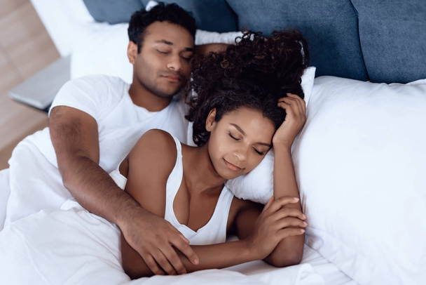 Μαύρος άνδρας και γυναίκα στην κρεβατοκάμαρα. Μια γυναίκα είναι ξαπλωμένη στο κρεβάτι και αγκαλιάζει τον άντρα της. Κοιμούνται Αγκαλιάζοντας κάθε άλλο. - Φωτογραφία, εικόνα